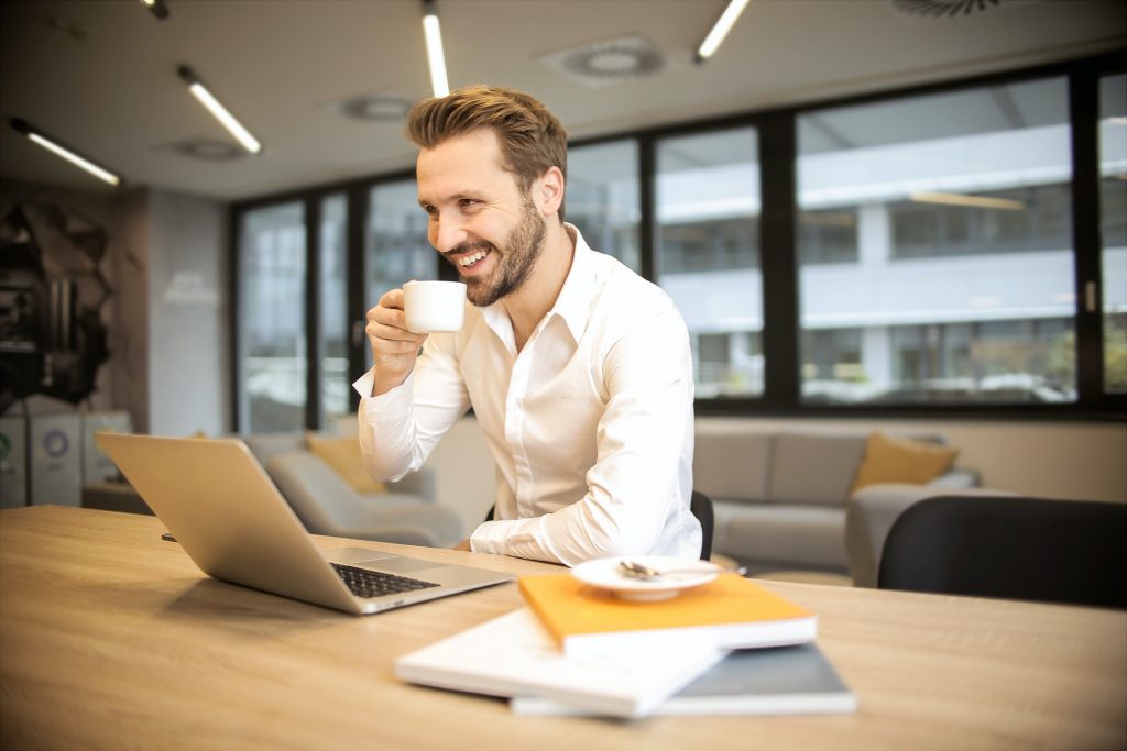 A smiling man drinks a cup of coffee from behind a laptop