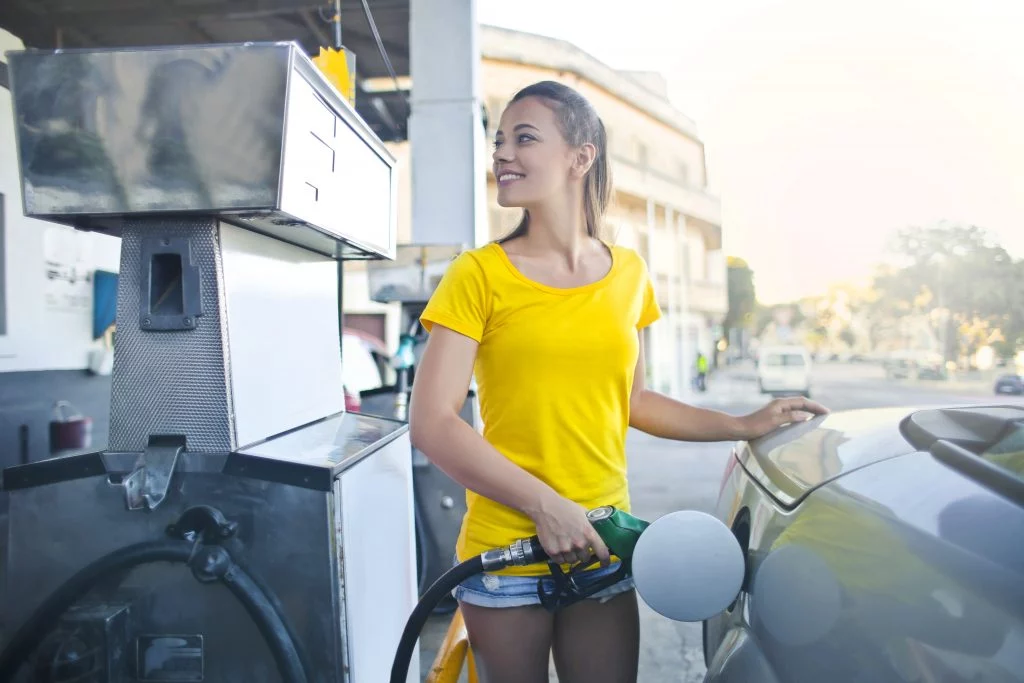 A smiling woman in a yellow tee-shirt fuels a silver car at a gas pump.