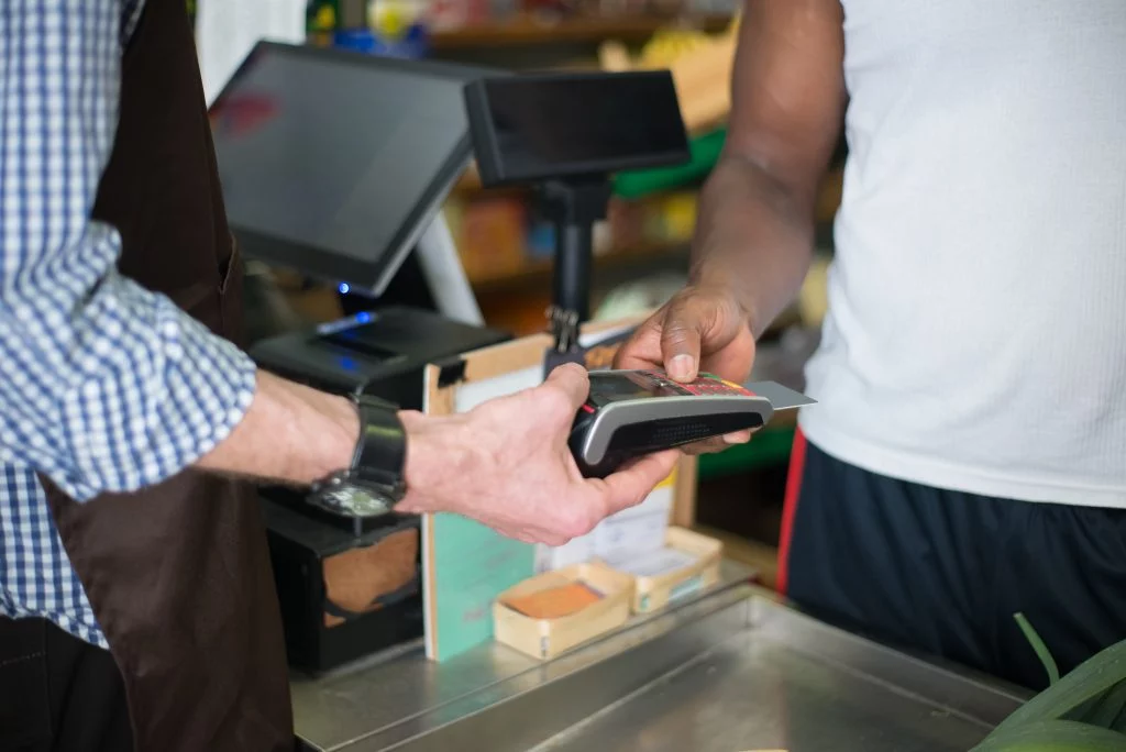 A man behind a checkout terminal holds a card reader, while another man is entering his pin number into the reader