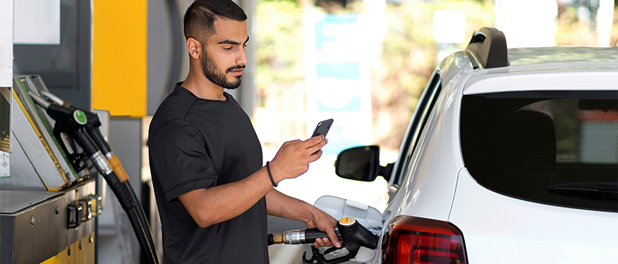 A man refuels a white SUV while looking at a mobile phone.